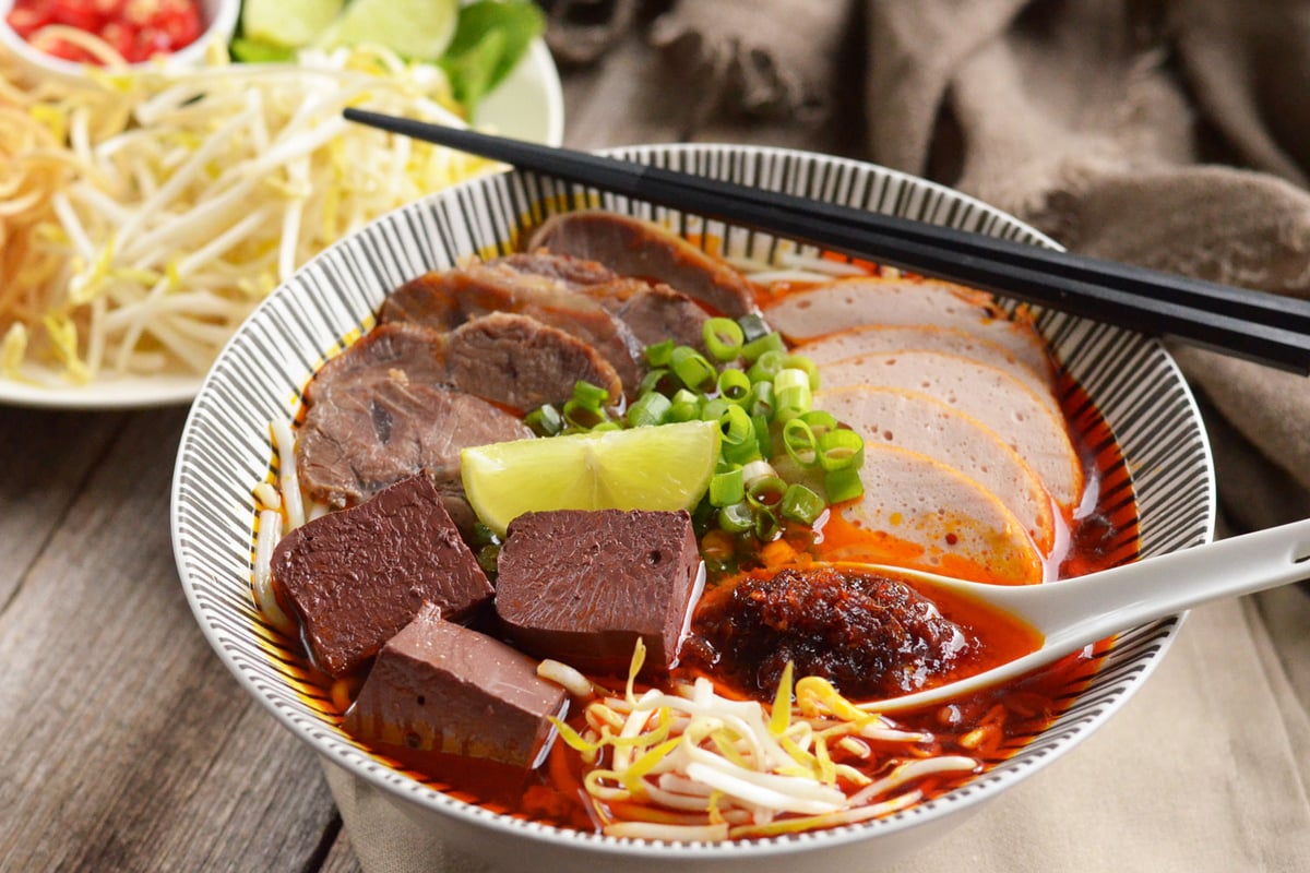 Bun Bo Hue is served with thick noodles, beef slices, and sometimes pork knuckle or blood pudding